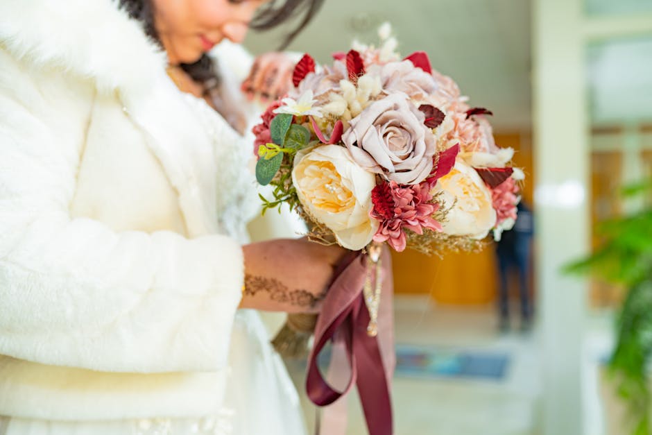A woman in a white, textured coat holds a large, round floral bouquet with soft pink, cream, and burgundy roses, complemented by green foliage. The bouquet is wrapped in elegant pink fabric ribbons, s