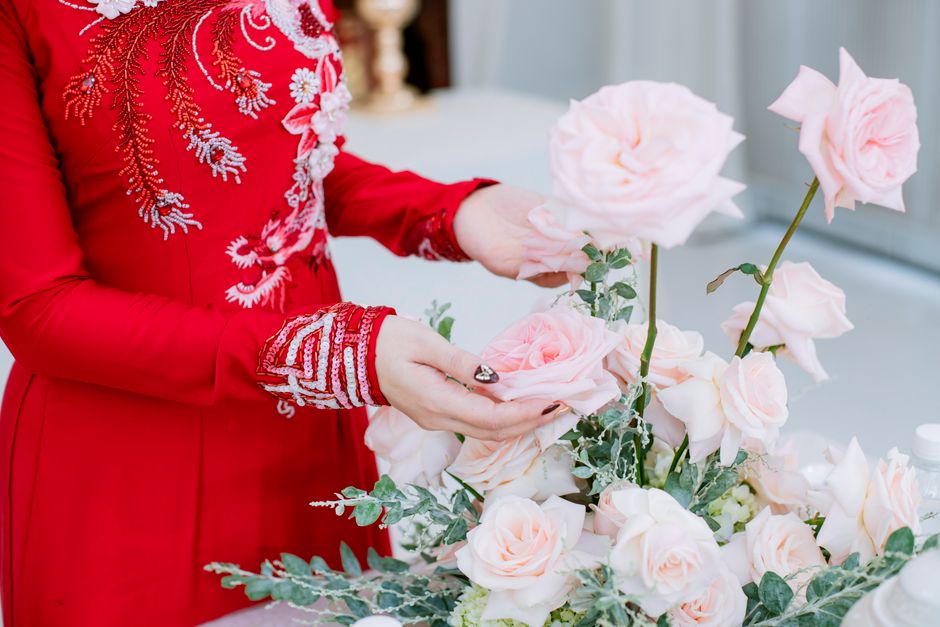 A person wearing a vibrant red dress with intricate white floral embroidery arranging a bouquet of large, pale pink roses with green foliage, using both hands for support and positioning the flowers o
