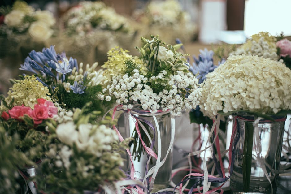 A collection of fresh floral bouquets arranged in clear glass vases, featuring predominantly white hydrangeas, delicate white baby's breath, pale pink roses, and light blue flowers. The flowers are ar