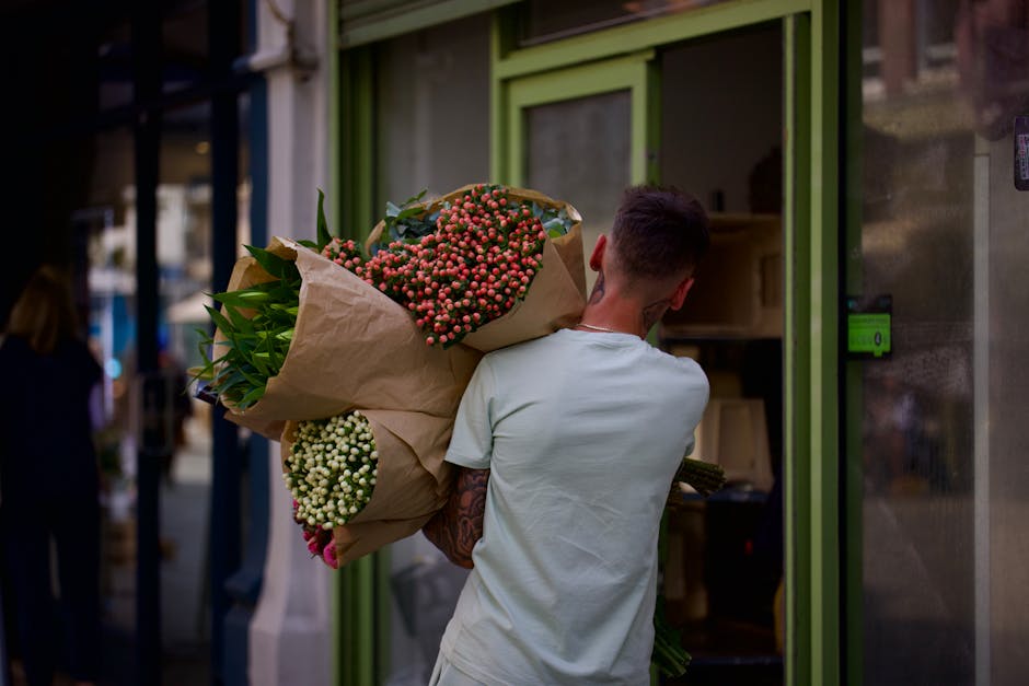 A person with short dark hair, dressed in a light-colored t-shirt, is seen from behind holding a large bouquet of fresh flowers wrapped in brown paper, featuring clusters of pink and white flowers wit