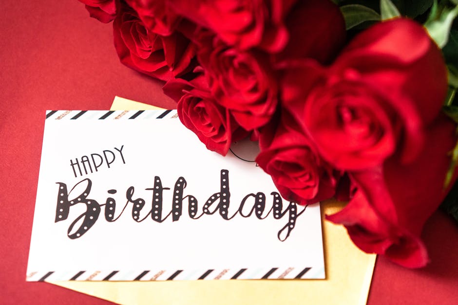 A vibrant bouquet of fresh red roses with tightly curled petals, arranged in a classical style, placed on a red surface. Some green foliage is visible among the roses, adding contrast and texture. The