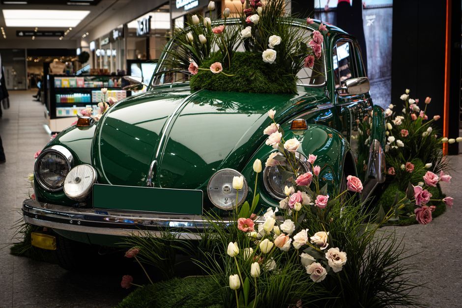 A vintage dark green Volkswagen Beetle decorated with a floral display featuring pink and white roses, tulips, and greenery, arranged on the hood, roof, and surrounding the base of the car. The flower
