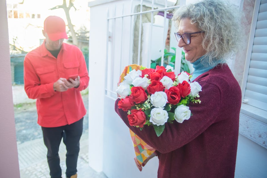 A woman with curly gray hair, glasses, and a burgundy cardigan holds a vibrant bouquet of fresh red and white roses wrapped in patterned paper, standing near a window in a well-lit, modern home enviro