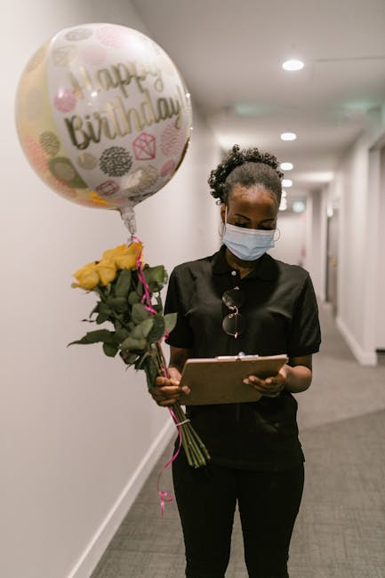 A young woman with dark curly hair, wearing a black uniform and a protective face mask, stands in a well-lit corridor holding a bouquet of yellow roses tied with a pink ribbon and a round 'Happy Birth