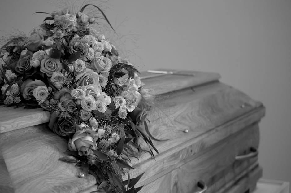A funeral floral arrangement resting on the lid of a wooden casket, featuring a lush bouquet of roses, carnations, and other flowers with varying sizes and textures. The flowers include both fully blo
